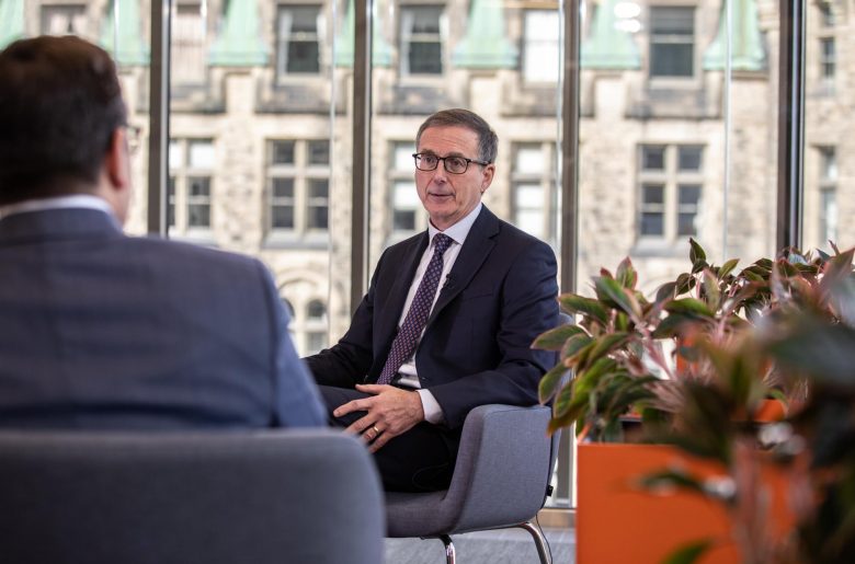 Tiff Macklem sitting comfortably in an easy chair. He is speaking to an interviewer, who is sitting with his back to the camera.