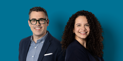 Two people smiling at the camera: a middle-aged man wearing glasses with fair skin and light brown hair that is grey on the sides, and a woman with medium skin and long curly dark hair.