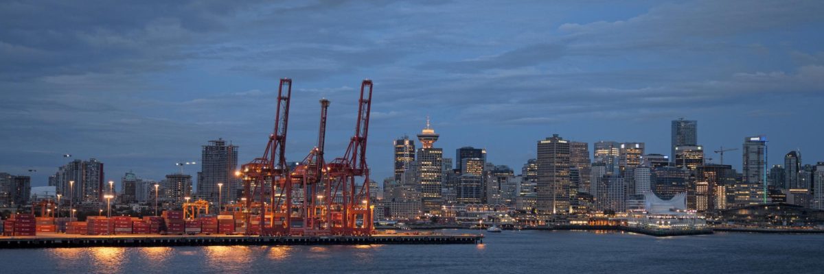Port of Vancouver, Canada Three large cranes can be seen against the skyline of east Vancouver. Many shipping containers can be seen directly behind the cranes.